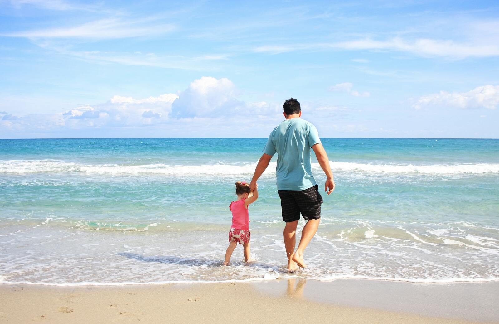 A father and daughter hold hands while walking on a sunny beach, enjoying family time together.
