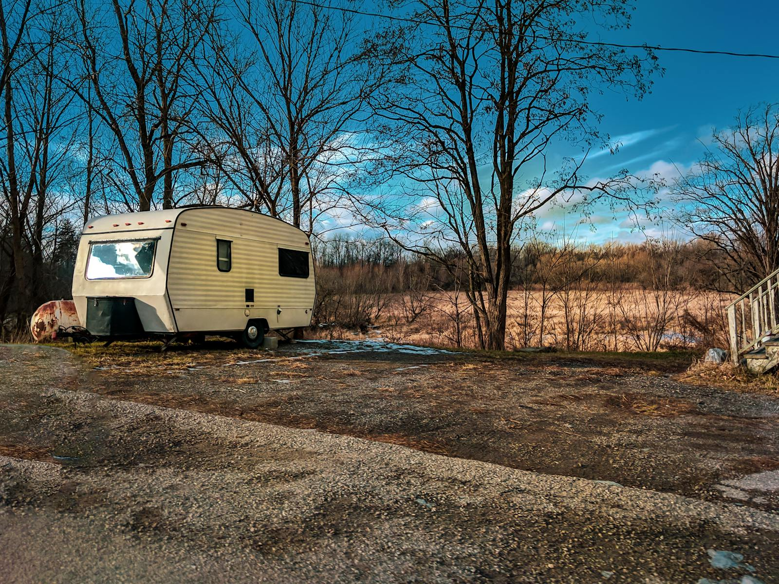 A lone trailer parked amidst bare trees and bare field under a cloudy blue sky.