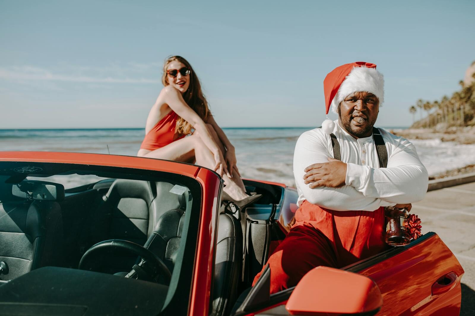 Santa Claus enjoying a holiday ride with a woman in a red convertible by the beach.