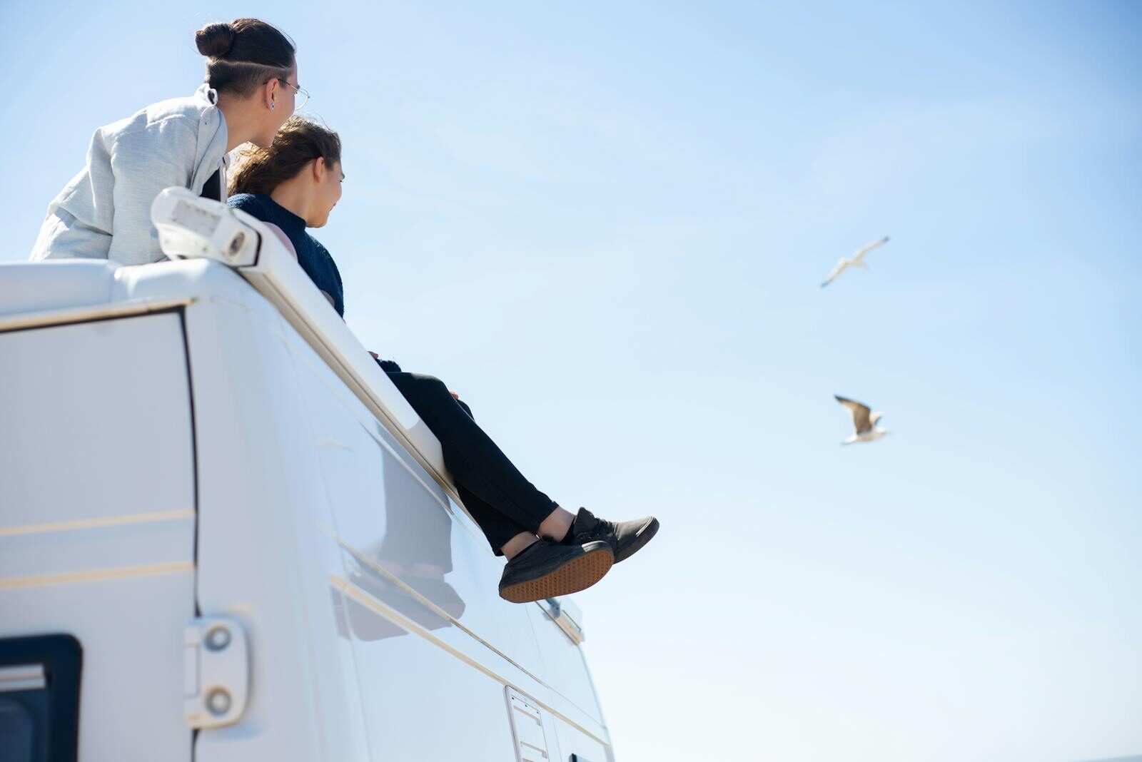 Two people sitting on a camper van roof enjoying a sunny day with seagulls flying.